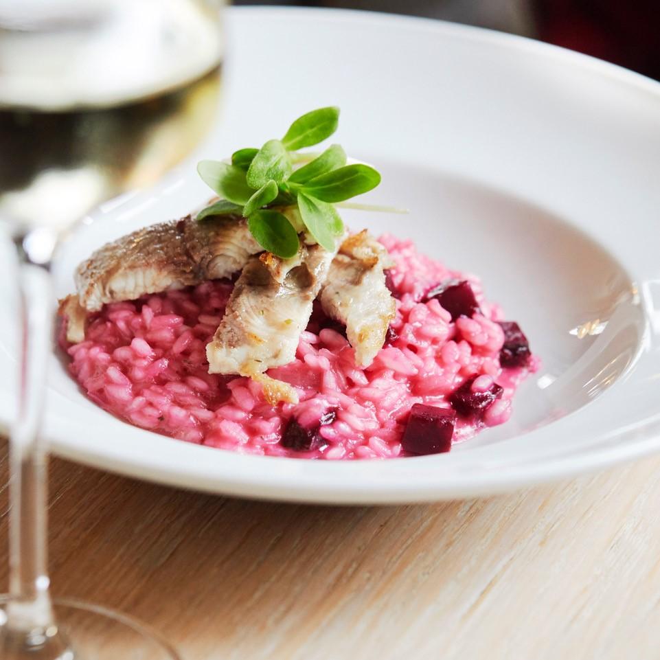 Grilled fish filet on pink beetroot risotto garnished with fresh herbs in white bowl.