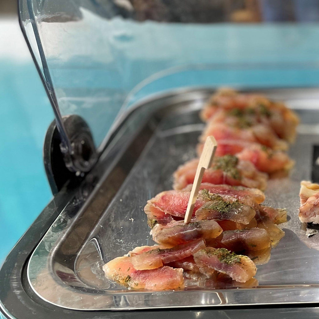 Thinly sliced pink fish fillet with herbs and seasonings on a silver tray with toothpicks.
